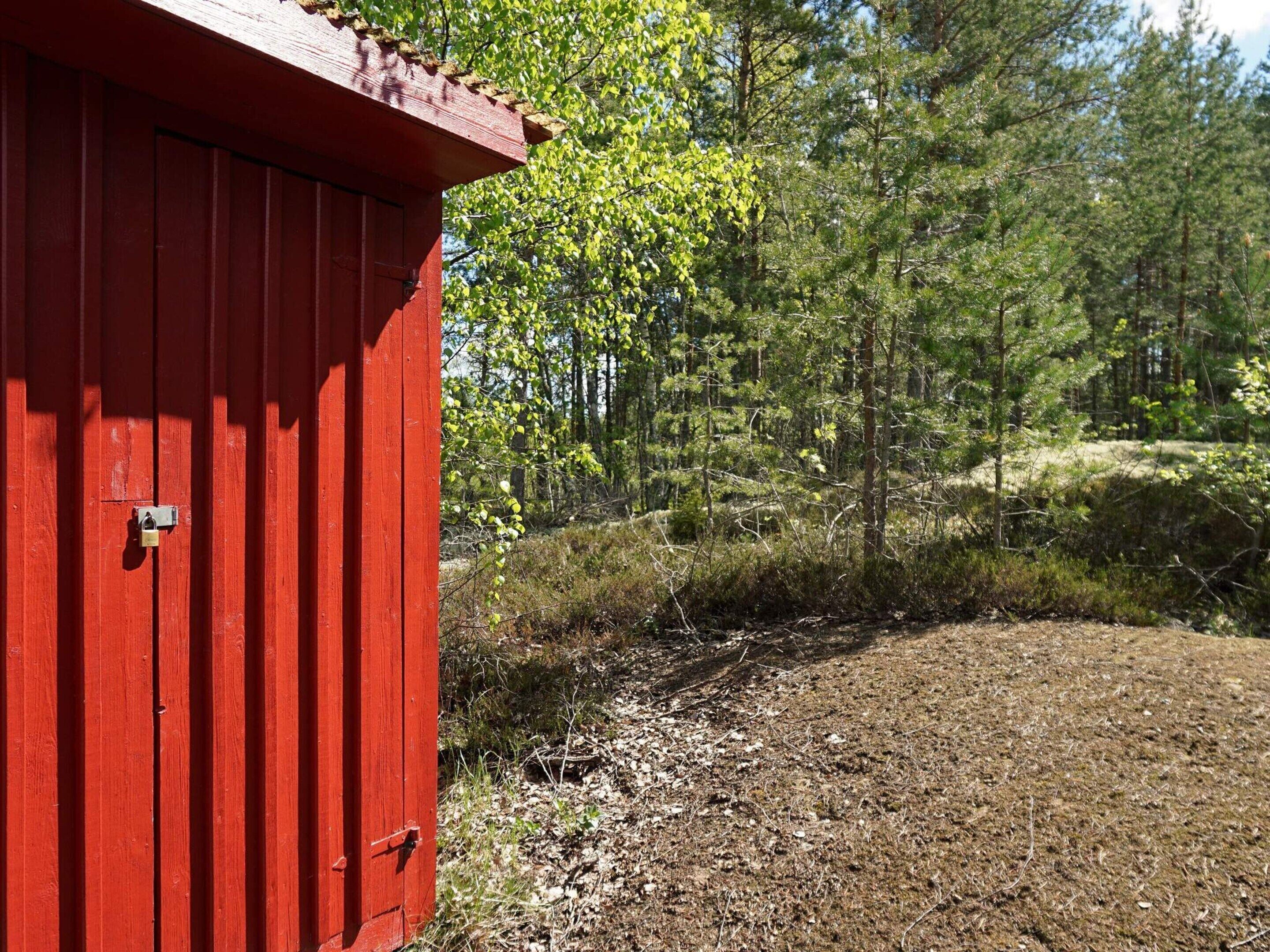 Una estructura roja de madera con una puerta de metal, rodeada por un área boscosa con árboles y matorrales.