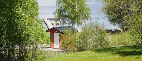 A small red building with a white door, surrounded by greenery and overlooking a calm body of water.