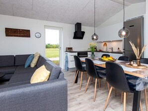 A modern living room with a grey sofa, a dining table with chairs, a kitchen area with a stove, and a large window.