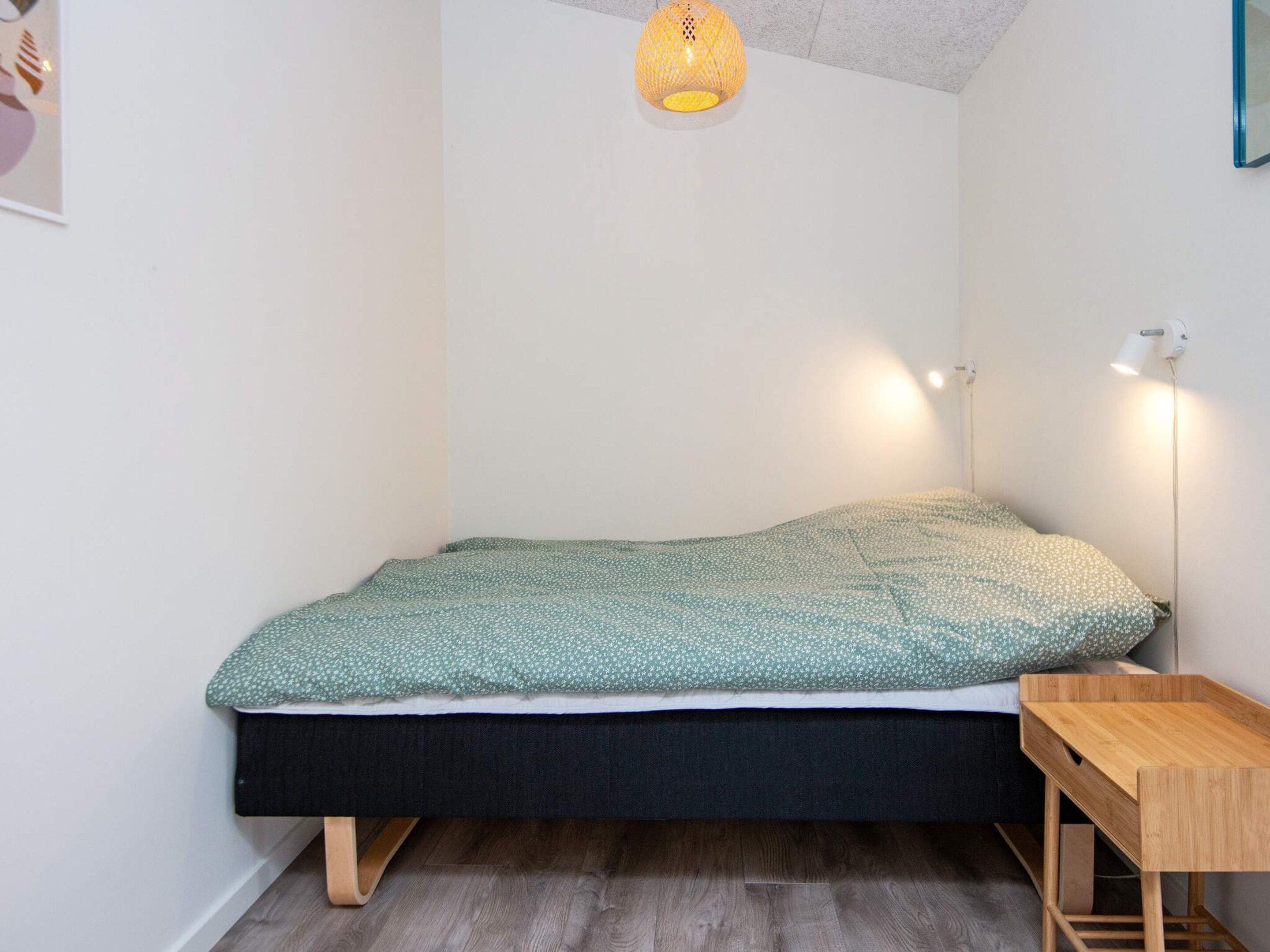 Een eenpersoonsbed met een bedspreider met groene patronen, een houten nachtkastje en een hanglamp.
