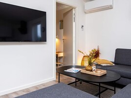 Una sala moderna con una televisión de pantalla plana montada en la pared, una mesa redonda de centro con una revista, un jarrón con flores y un sofá gris.