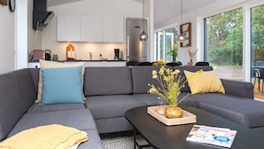 A modern living room with a grey sectional sofa, a coffee table with a magazine, and a kitchen in the background. - 5 Star Holiday Home in Ansager (Ansager)