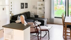 A modern living space with a kitchen area, a dining table, and a sofa. There is a view of a garden through a large window.