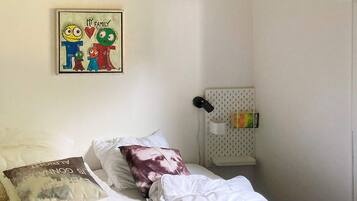 A bedroom with a bed, pillows, a wall-mounted shelf with a box, and a framed picture on the wall.