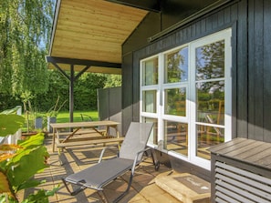 Ein Terrassenbereich im Freien mit einem Holztisch und -stühlen, einer Liege und einem großen Fenster mit vielen Scheiben.