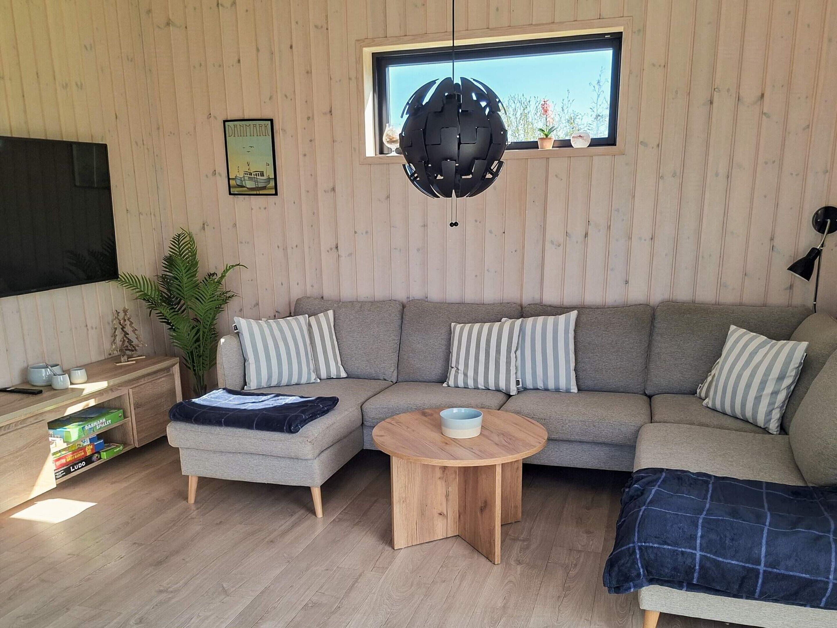 A modern living room with a sectional sofa, a round wooden coffee table, a TV on the left, and a window with a view of plants.