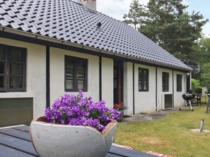 Ett hus med ett tak täckt av tegelpannor och vita väggar, en blomkruka med lila blommor på ett bord och en grill i trädgården.