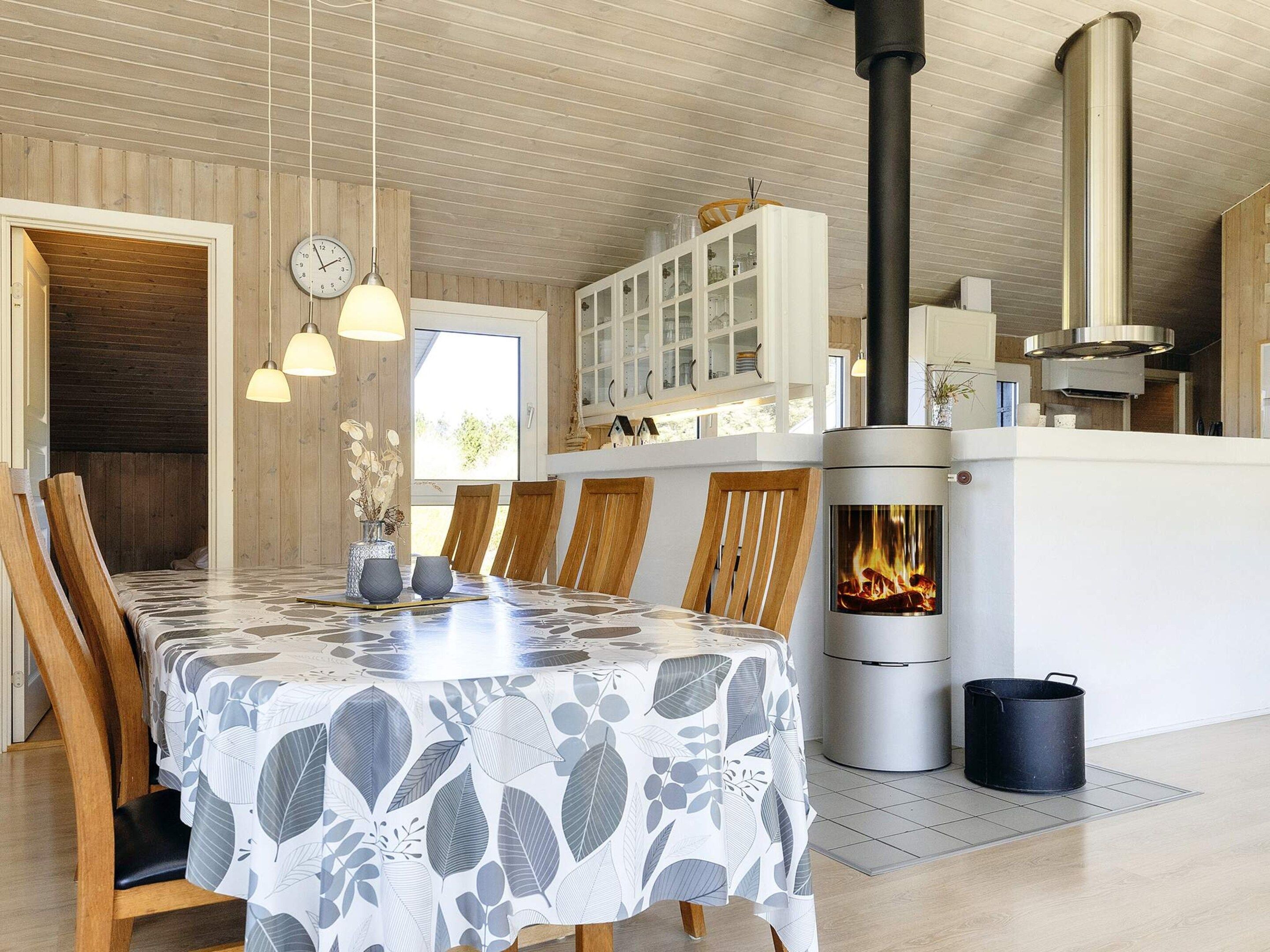 A dining area with a table covered in a leaf-patterned tablecloth, wooden chairs, pendant lights, a stove with a visible fire, and a white cabinet with glass doors.