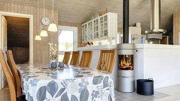 A dining area with a table covered in a leaf-patterned tablecloth, wooden chairs, pendant lights, a stove with a visible fire, and a white cabinet with glass doors.