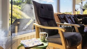 A cozy seating area with a leather chair and a cushioned bench, a table with a mug and a magazine, and a view of greenery through a large window.
