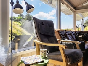 A cozy seating area with a leather chair and a cushioned bench, a table with a mug and a magazine, and a view of greenery through a large window. - 14 Person Holiday Home in Blokhus (Blokhus)