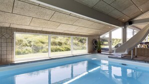 An indoor swimming pool with clear glass walls, a slide, and a view of greenery outside.