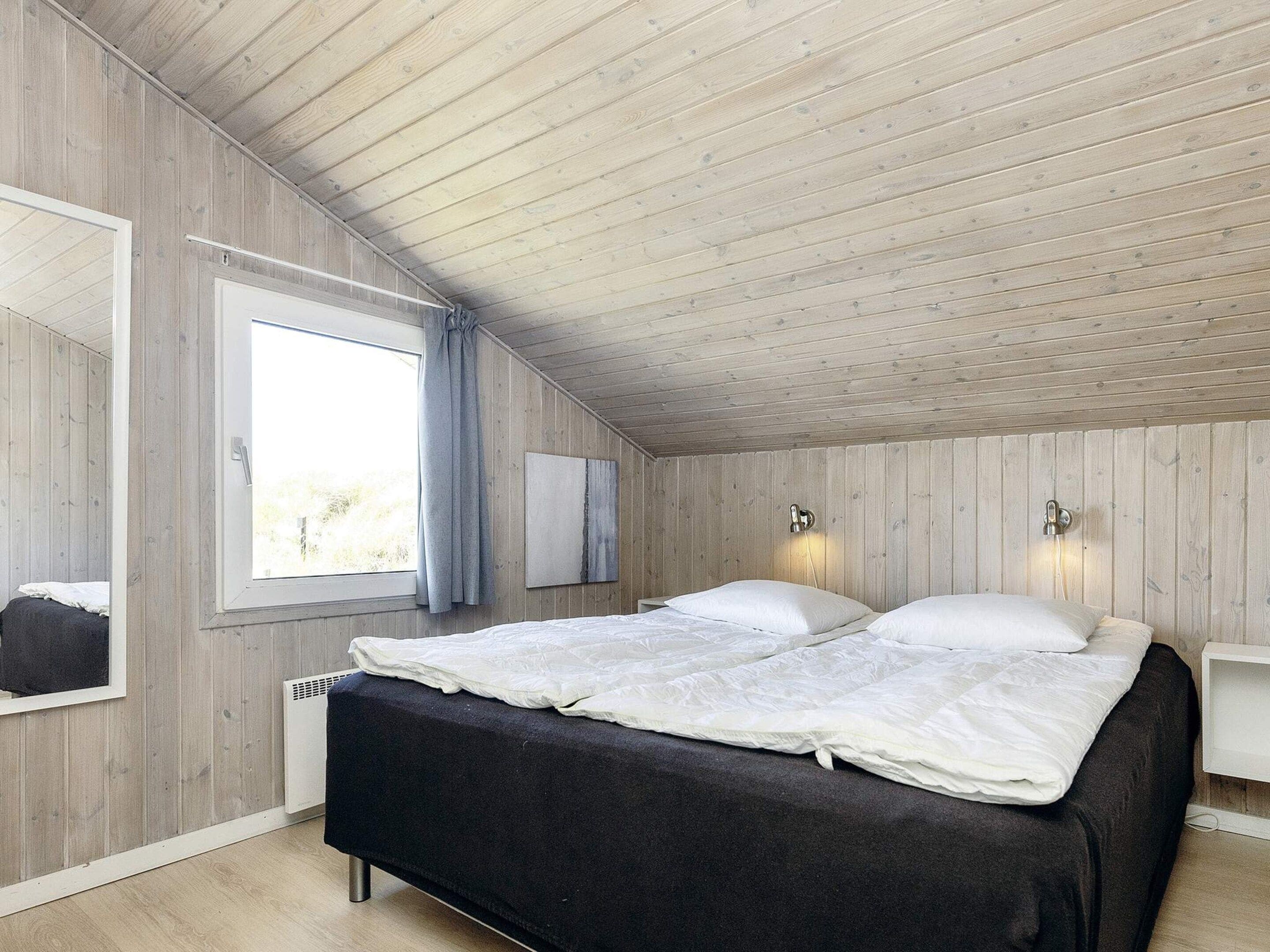 Ein Schlafzimmer mit Holzboden, einem Bett mit weißer Bettwäsche, einem Fenster mit Vorhängen und einer wandmontierten Lampe.