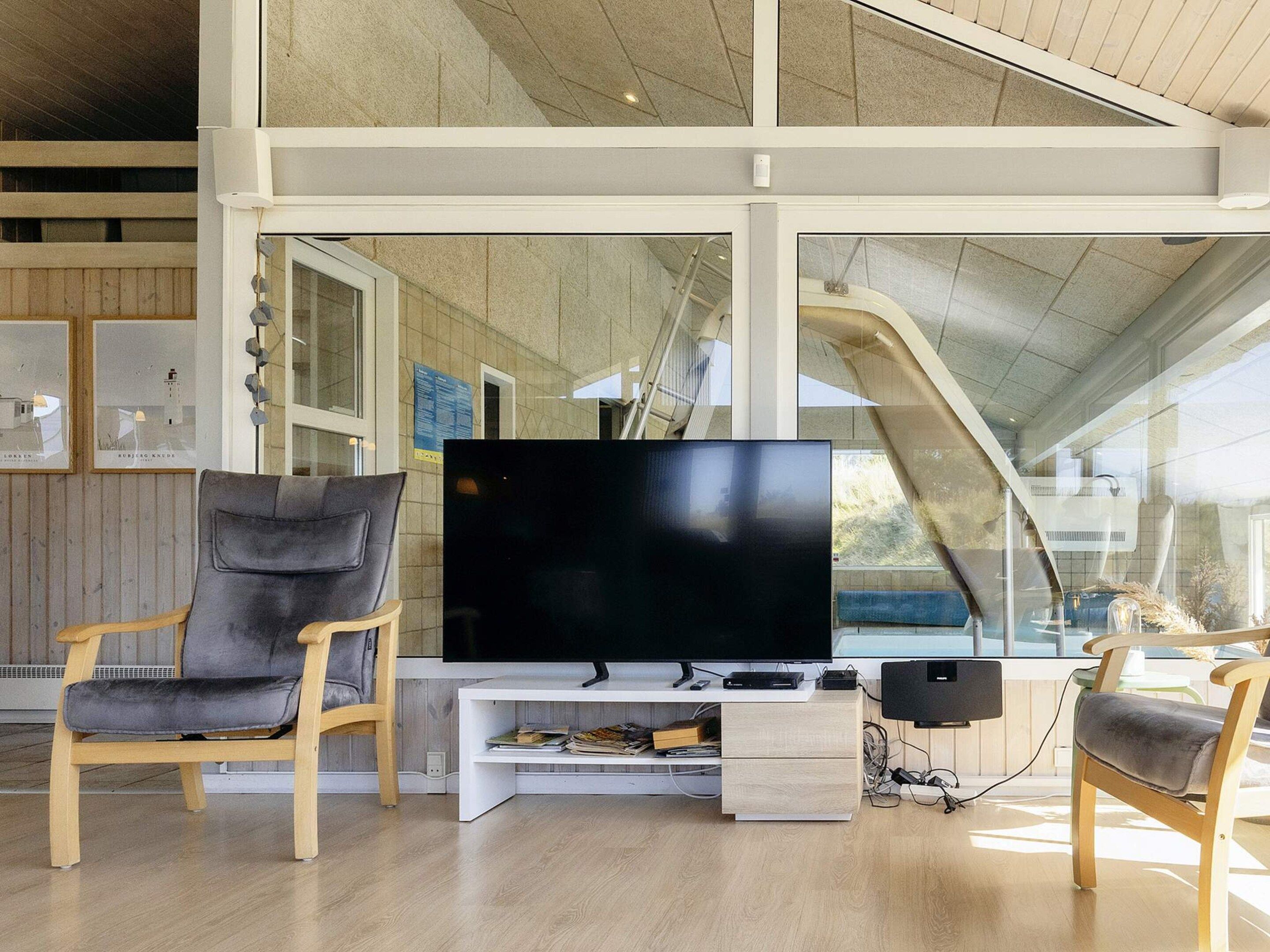 A modern living room with a large flat-screen TV, wooden chairs, a white entertainment unit, and a view of a building with a sloped roof.