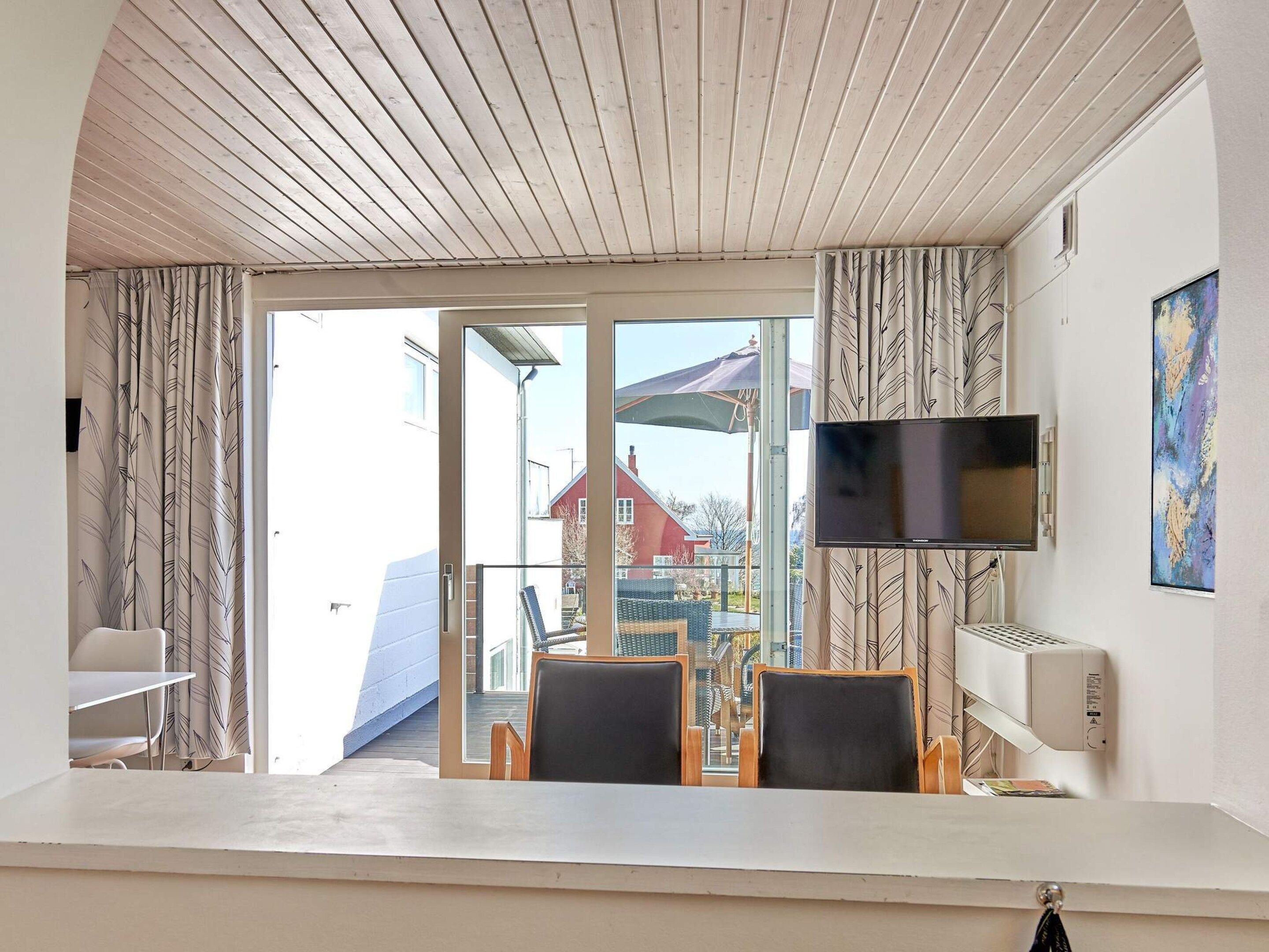 A room with a wooden ceiling, a TV mounted on the wall, a table with chairs, and a view of the outside through a glass door.
