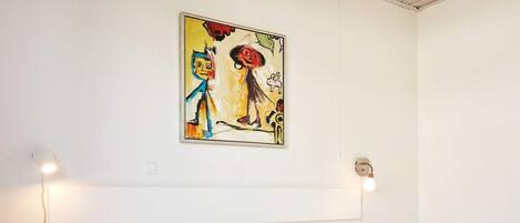 A bedroom with a bed, a nightstand, a wall-mounted painting, and two wall-mounted lights.