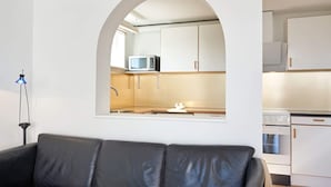 Una sala de estar moderna con un sofá de cuero negro, una cocina visible a través de un ventanal arqueado y una lámpara de pie azul.