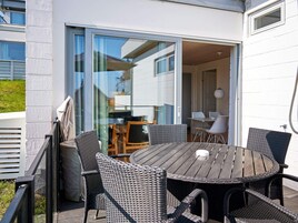 Un patio exterior con una mesa redonda y sillas de mimbre, una puerta corrediza de vidrio que da a un espacio interior y una pared exterior blanca.