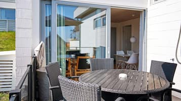 Un patio exterior con una mesa redonda y sillas de mimbre, una puerta corrediza de vidrio que da a un espacio interior, y una pared exterior blanca.