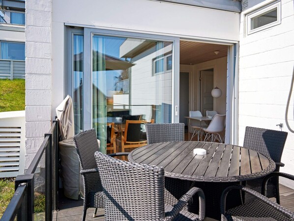 An outdoor patio with a round table and wicker chairs, a sliding glass door leading to an interior space, and a white exterior wall.