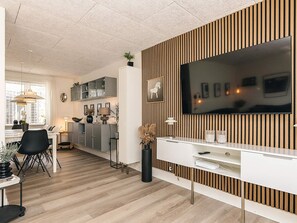 Un salon moderne doté d’un téléviseur à écran plat, d’un mur recouvert de panneaux en bois et d’un coin repas avec un buffet et des chaises blancs.