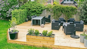 Eine Holzterrasse mit Gartenmöbeln, einem erhöhten Blumenbeet mit Pflanzen und einem Topfpflanzen auf dem Boden.