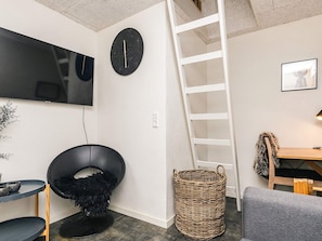 Une chambre moderne équipée d’un téléviseur à écran plat, d’un fauteuil noir, d’un panier en osier, d’une échelle en bois et d’un canapé gris.