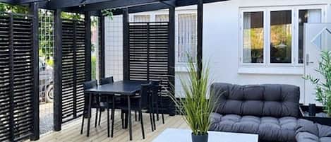 En overdækket udendørs siddeplads med et bord, stole og en polstret bænk, samt en potteplante.