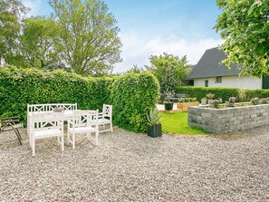 A gravel patio with a white wooden table and chairs, a hedge, and a house with a dark roof in the background.