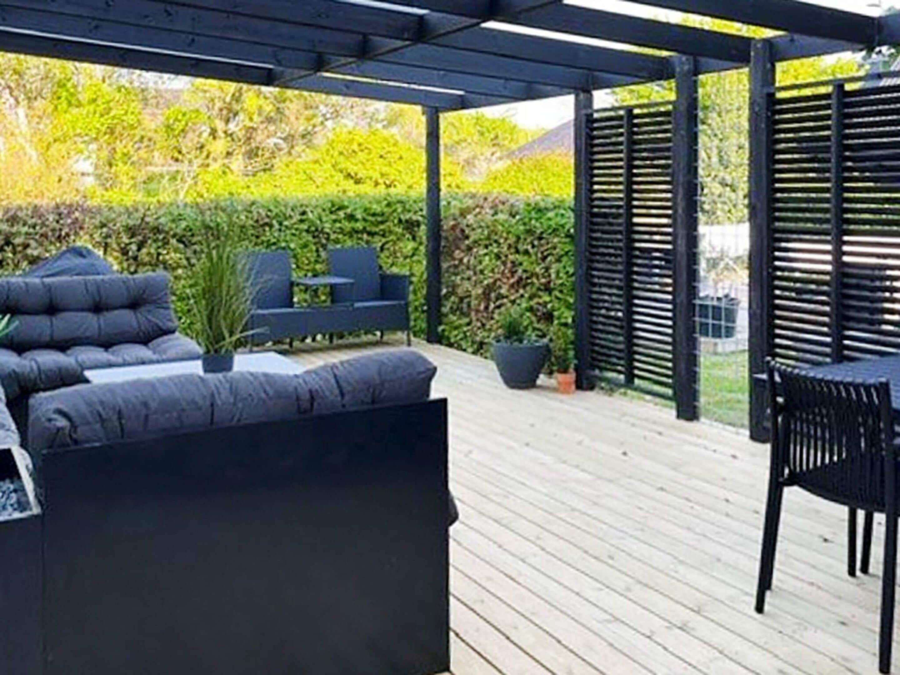 Un espace extérieur aménagé avec des sièges, abrité par un toit, comprenant un canapé et des fauteuils de couleur foncée, une petite table et des plantes en pot.