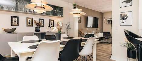 A modern dining area with a white table and black and white chairs, a TV mounted on the wall, and framed artwork on the walls.