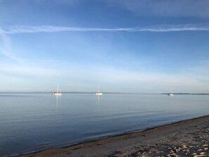 Mar tranquila con tres veleros anclados en la distancia, una playa arenosa en primer plano y un cielo despejado arriba.