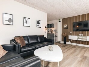Un salon moderne avec un canapé en cuir noir, une table basse blanche et un téléviseur à écran plat fixé à un mur en bois.