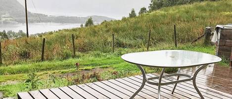 Eine Holzdielen-Terrasse mit rundem Tisch und Metallbeinen, mit Blick auf eine Rasenfläche mit Zaun und Bäumen.
