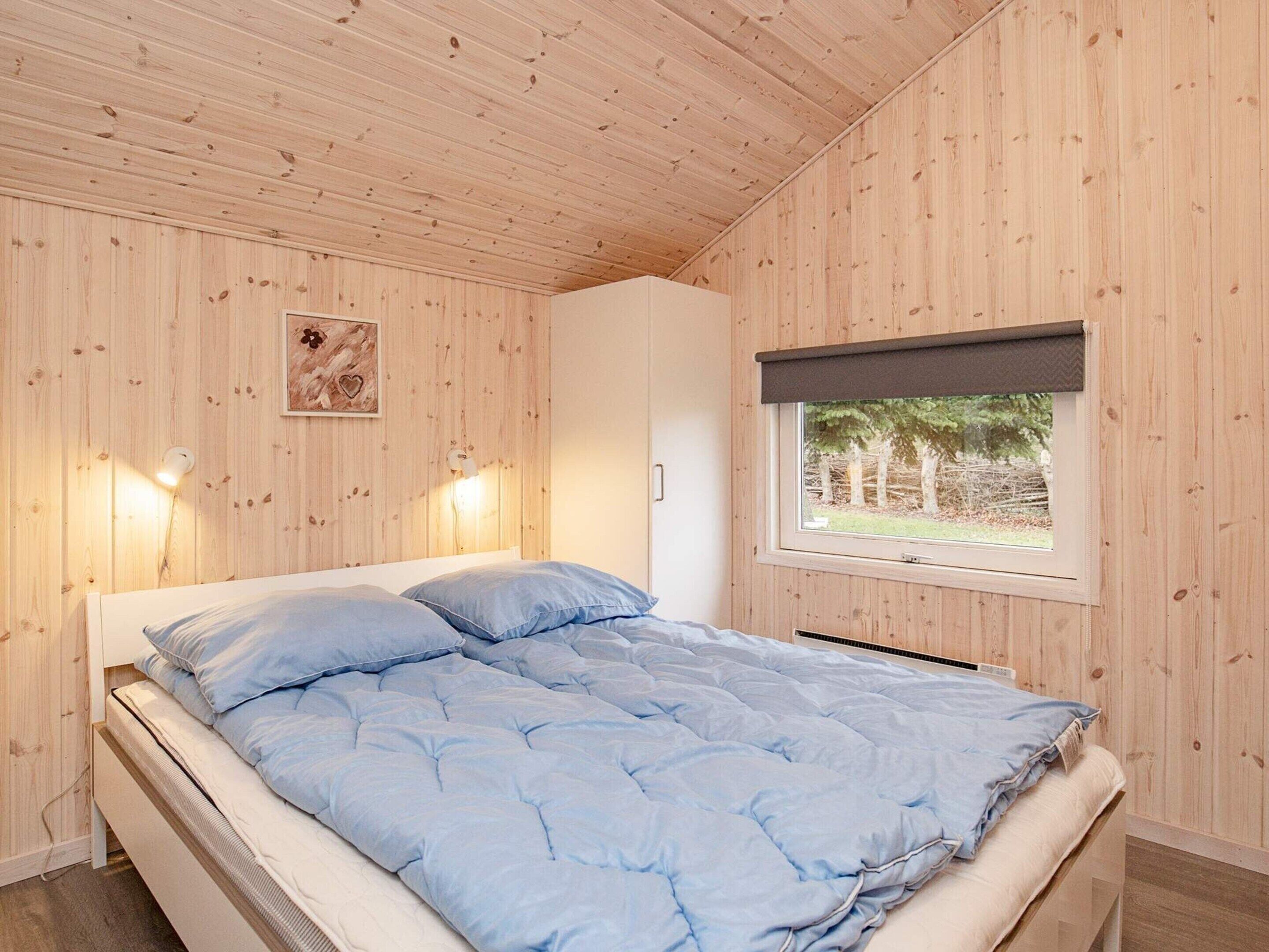 Un dormitorio con cama, paredes de madera, una ventana con vista a árboles y dos luces fijadas a la pared.