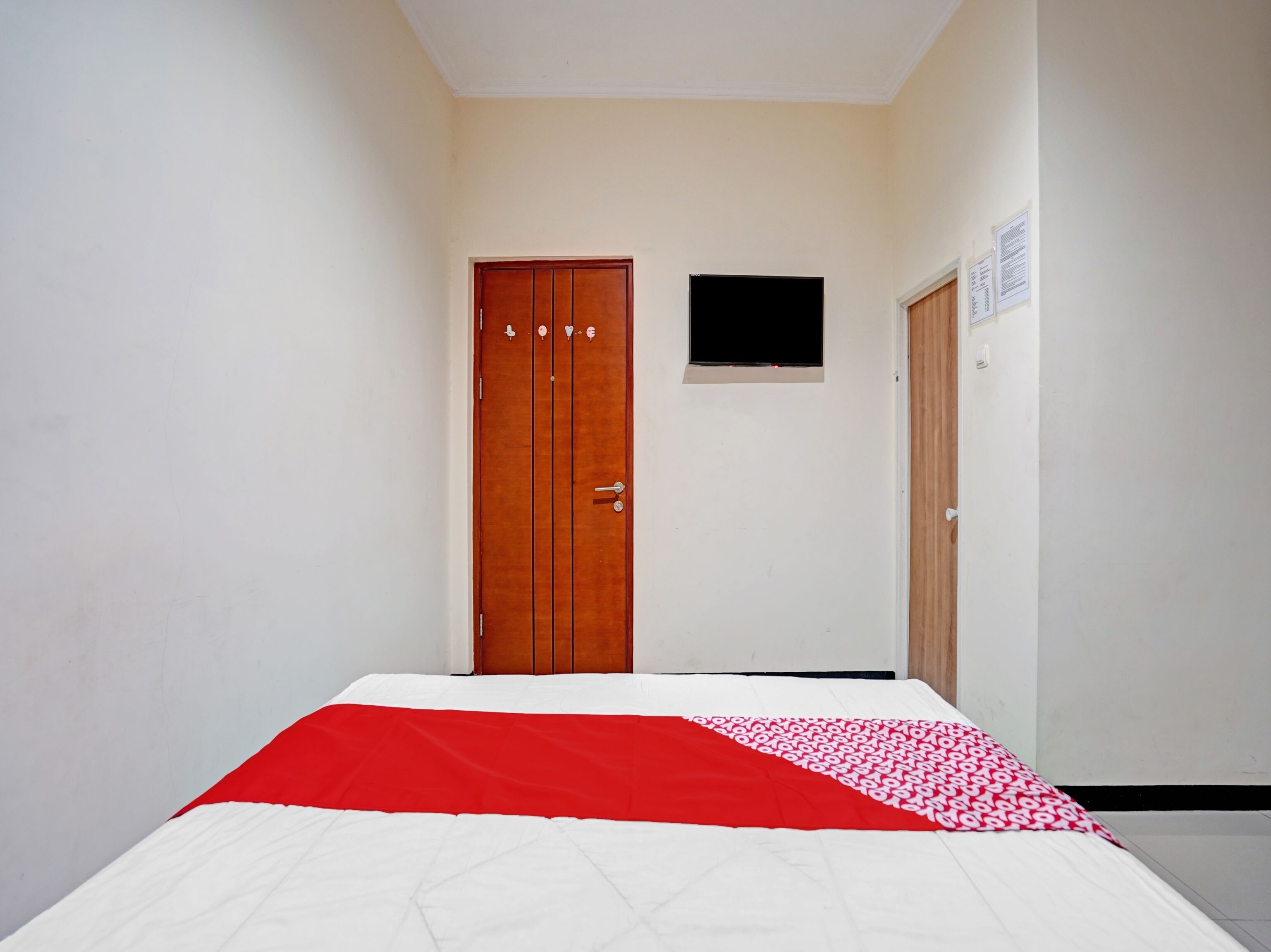 Une pièce avec un lit, une porte en bois, une petite fenêtre et un téléviseur fixé au mur.