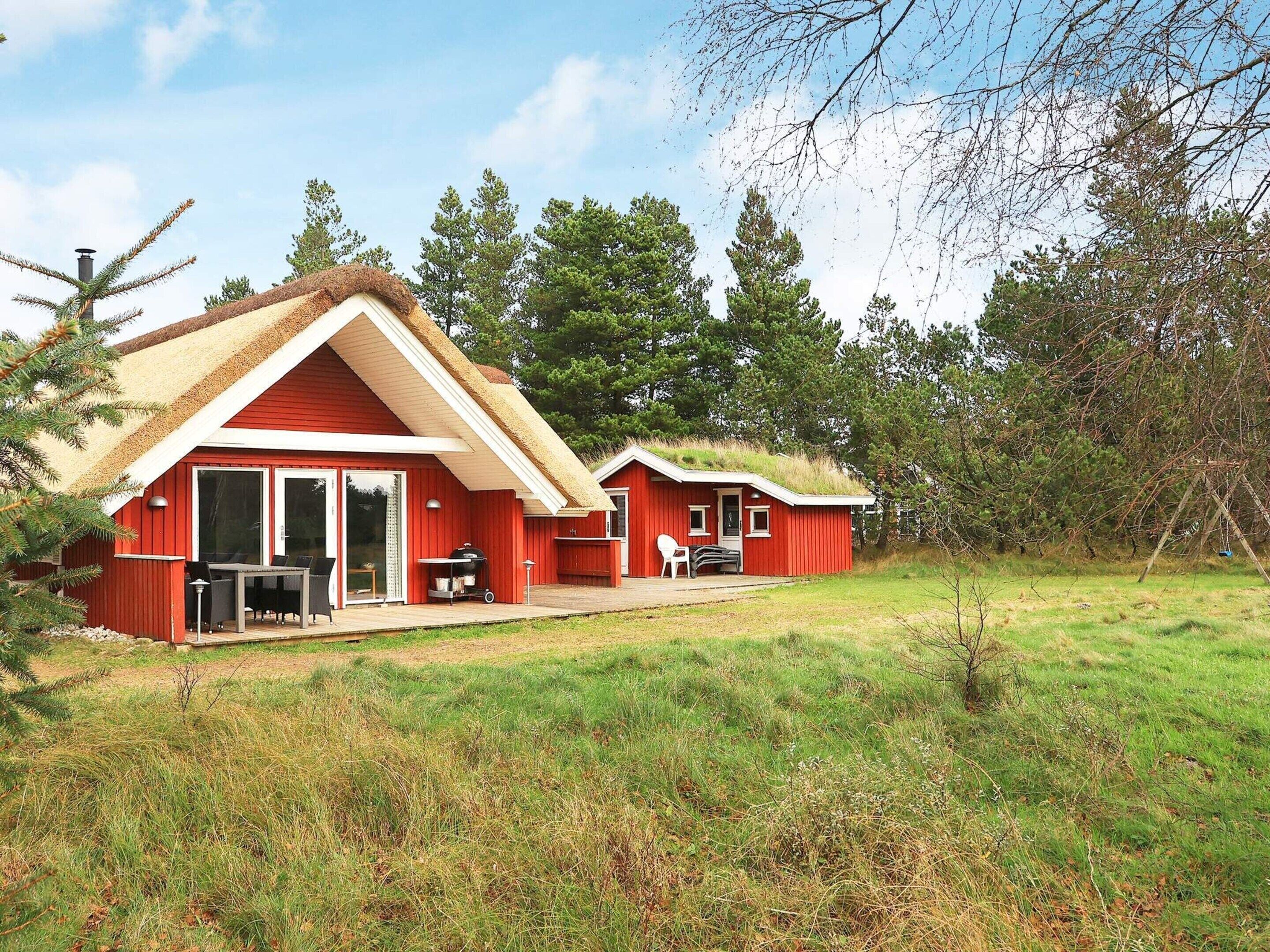 Une cabane rouge avec un toit de chaume, une terrasse avec des meubles d’extérieur et, au fond, un champ d’herbe.