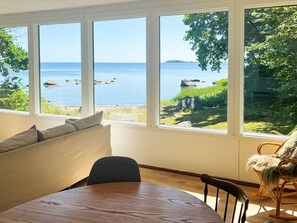 A room with a large window overlooking a body of water, a wooden table, a sofa, and a rocking chair. - 4 Star Holiday Home in Solvesborg (Sölvesborg)
