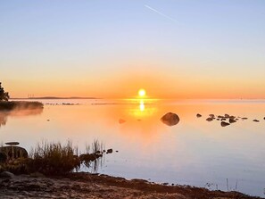 A calm body of water with a sunset, rocks, and a distant shoreline.