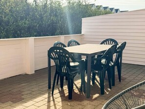 Um pátio com uma mesa de madeira e cadeiras pretas, cercado por uma parede branca e uma sebe.