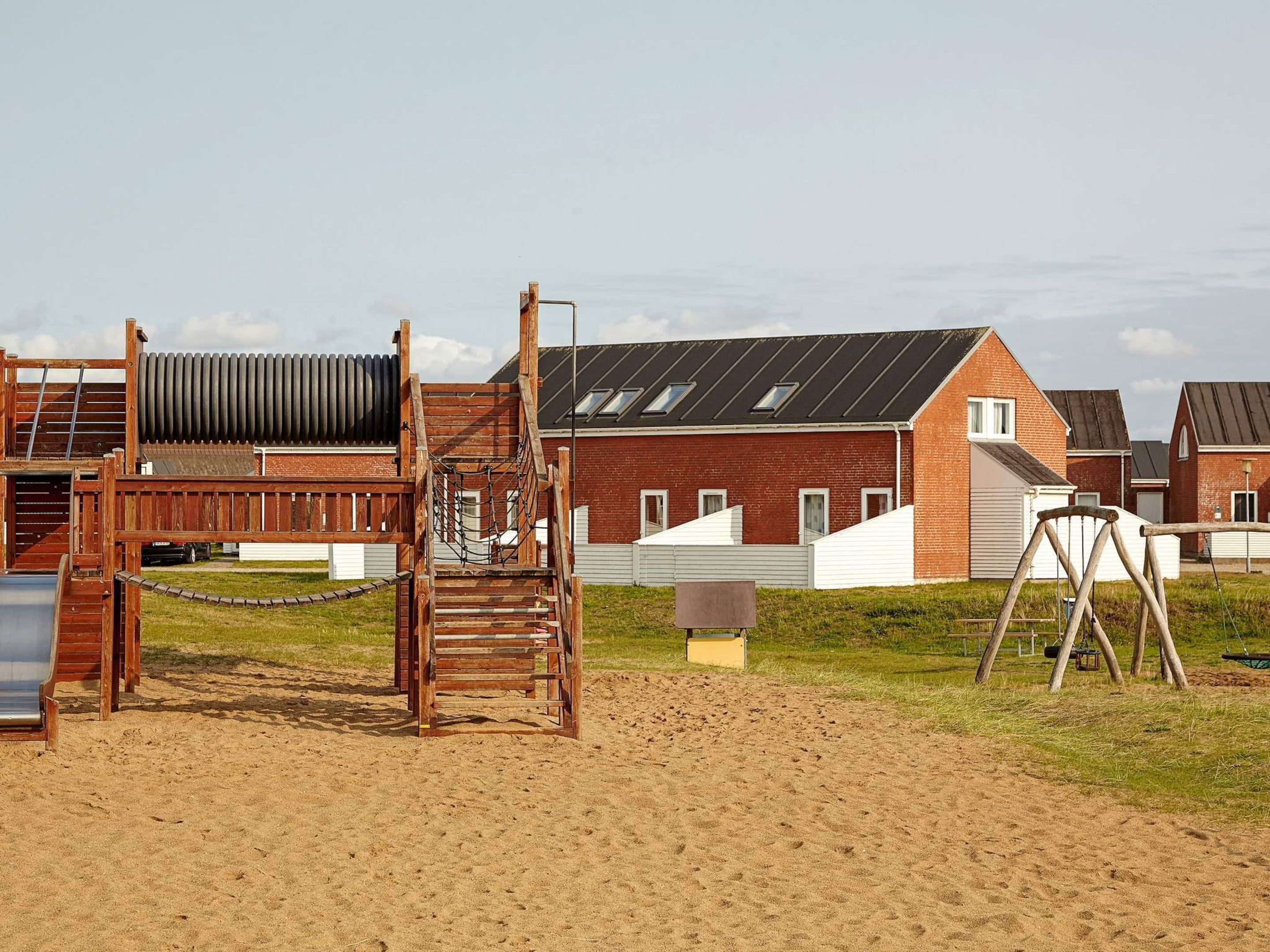 A playground with a wooden climbing structure and swings, set against a backdrop of residential houses with brick facades and sloped roofs.