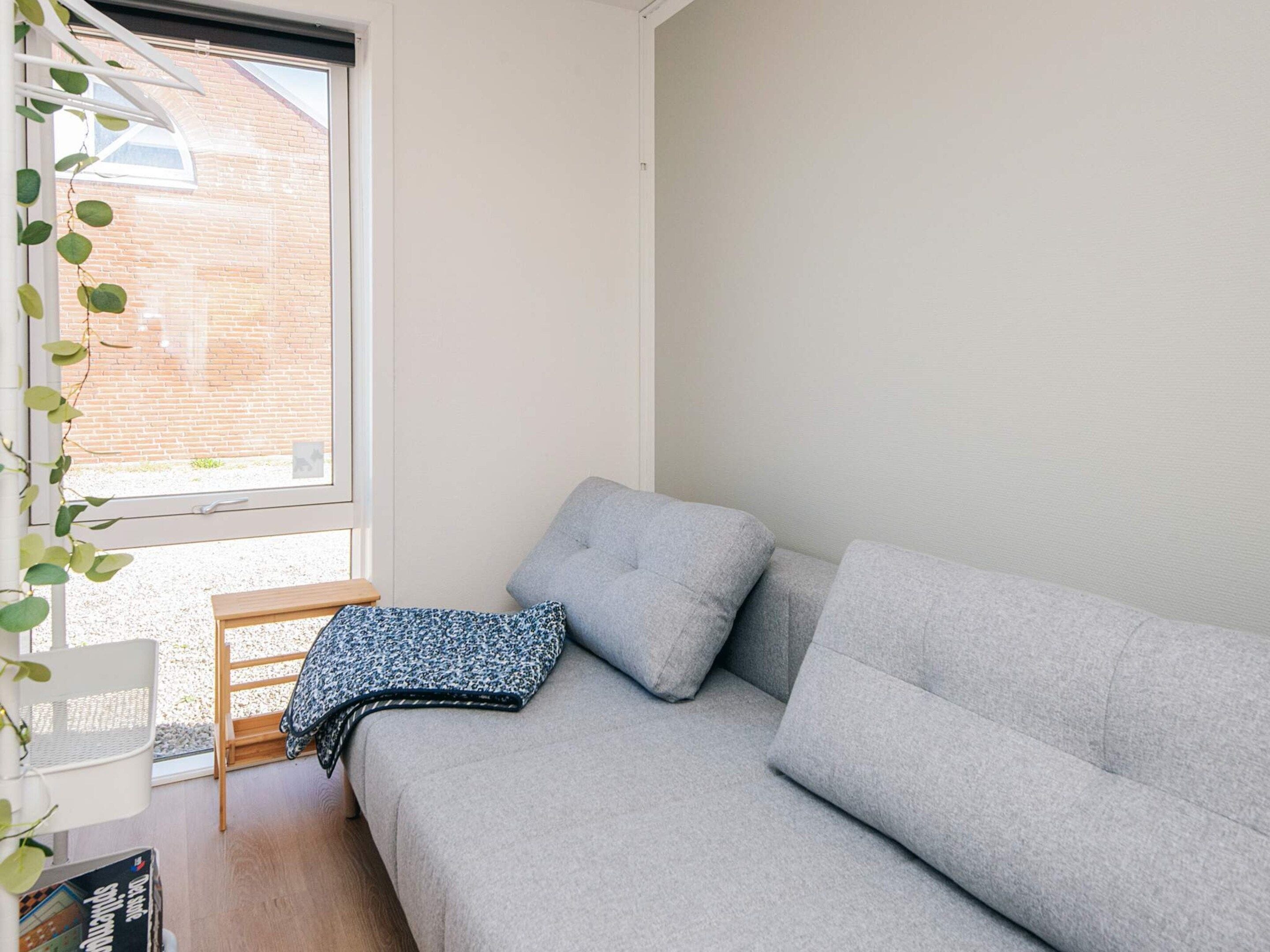 A corner room with a grey sofa, a small wooden chair, and a window with a view of a brick building.