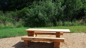 Una mesa de picnic de madera con un banco, ubicada en una zona de grava con césped y arbustos al fondo.