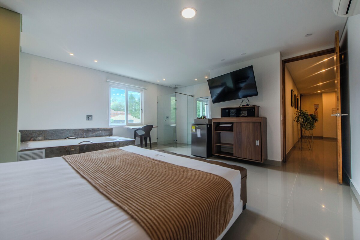 A modern hotel room with a large bed, a flat-screen TV, a minibar, and a glass-enclosed bathroom.