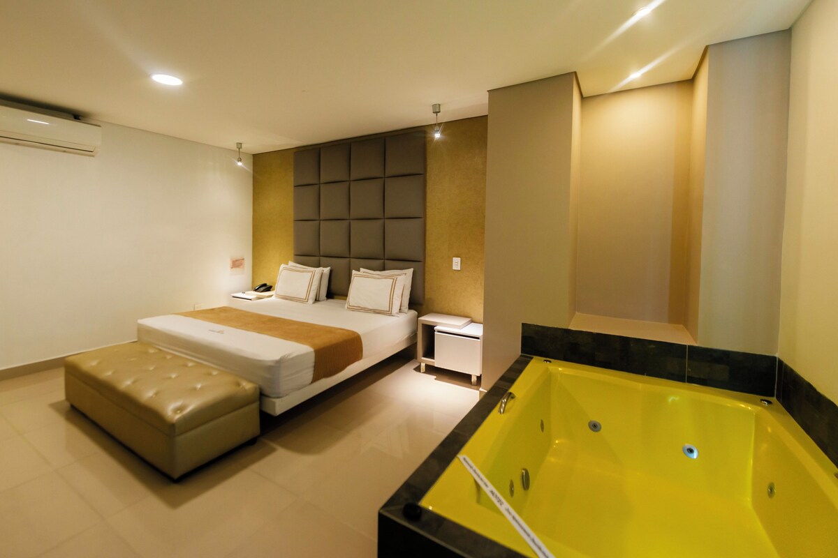 A modern hotel room with a large bed, a small bedside table, a bedside lamp, a built-in bench, and a yellow bathtub.