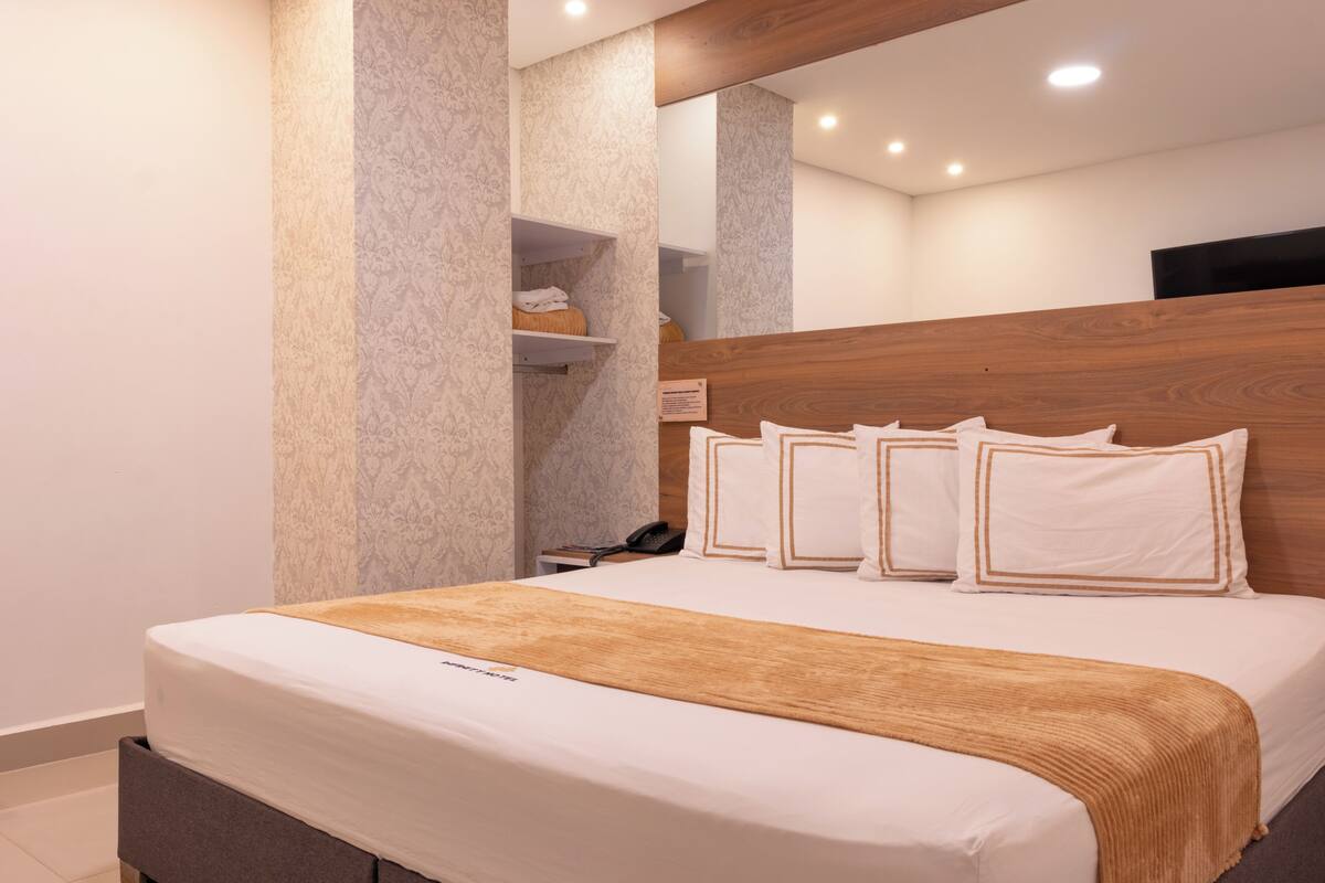 A modern hotel room with a wooden headboard, a large bed with white bedding and decorative pillows, a bedside table with a phone, and a built-in shelf with toiletries.