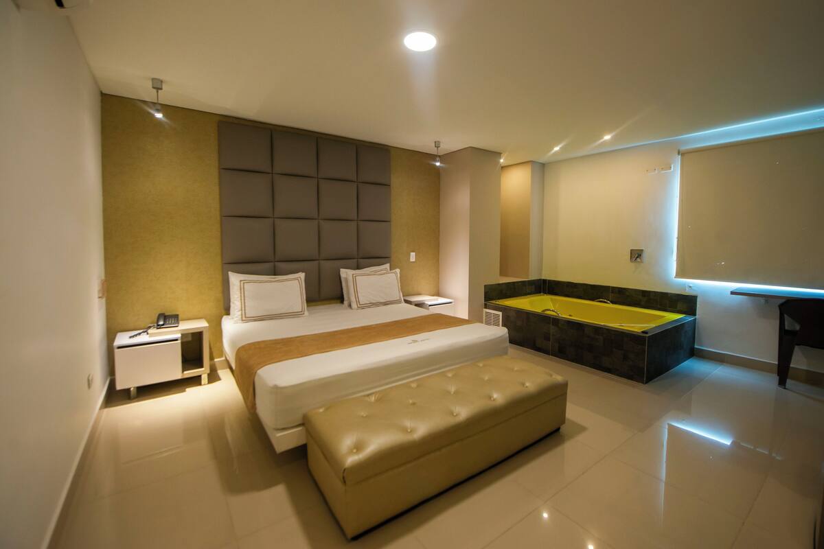 A modern hotel room with a large bed, a bench, a bedside table, a nightstand, a built-in bathtub, and a large window.