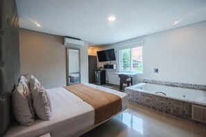 A hotel room with a bed, a large window, a TV, a desk, and a bathtub. - Infinity Hotel (Medellín)