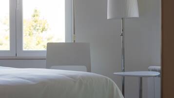 Habitación de hotel con cama, lámpara de pie, una mesita y ventana con vistas a edificios y árboles.