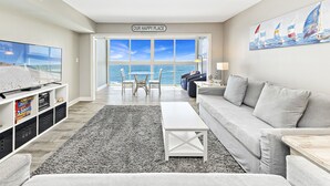 Un salon doté d’une grande fenêtre donnant sur la mer, d’un canapé blanc, d’une table basse et d’un téléviseur posé sur un meuble.
